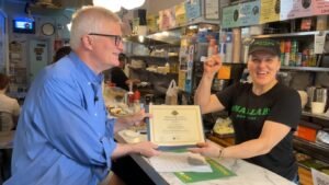 New York State Senator Brian Kavanagh presenting B&H Dairy NYS Historic Business Preservation Registry Award to a smiling woman behind the counter.
